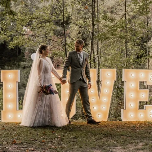 Brautpaar hält sich an den Händen vor großen leuchtenden „LOVE“-Buchstaben im Wald, romantische Outdoor-Hochzeit mit warmem Licht