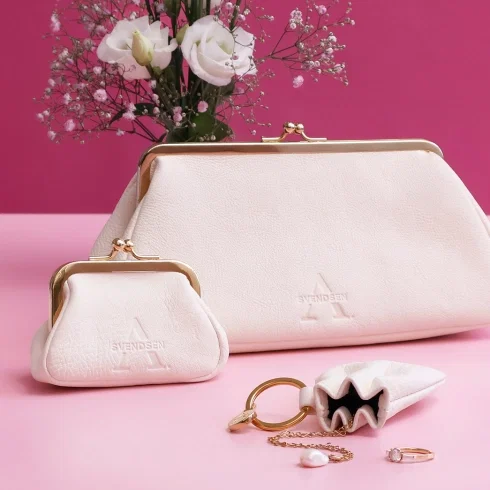 Elegantes Taschen-Set von Alexandra Svendsen mit weißer Abendtasche, kleinem Schmucktäschchen und Leder-Accessoire vor pinkem Hintergrund, dekoriert mit weißen Blumen und Schmuck.