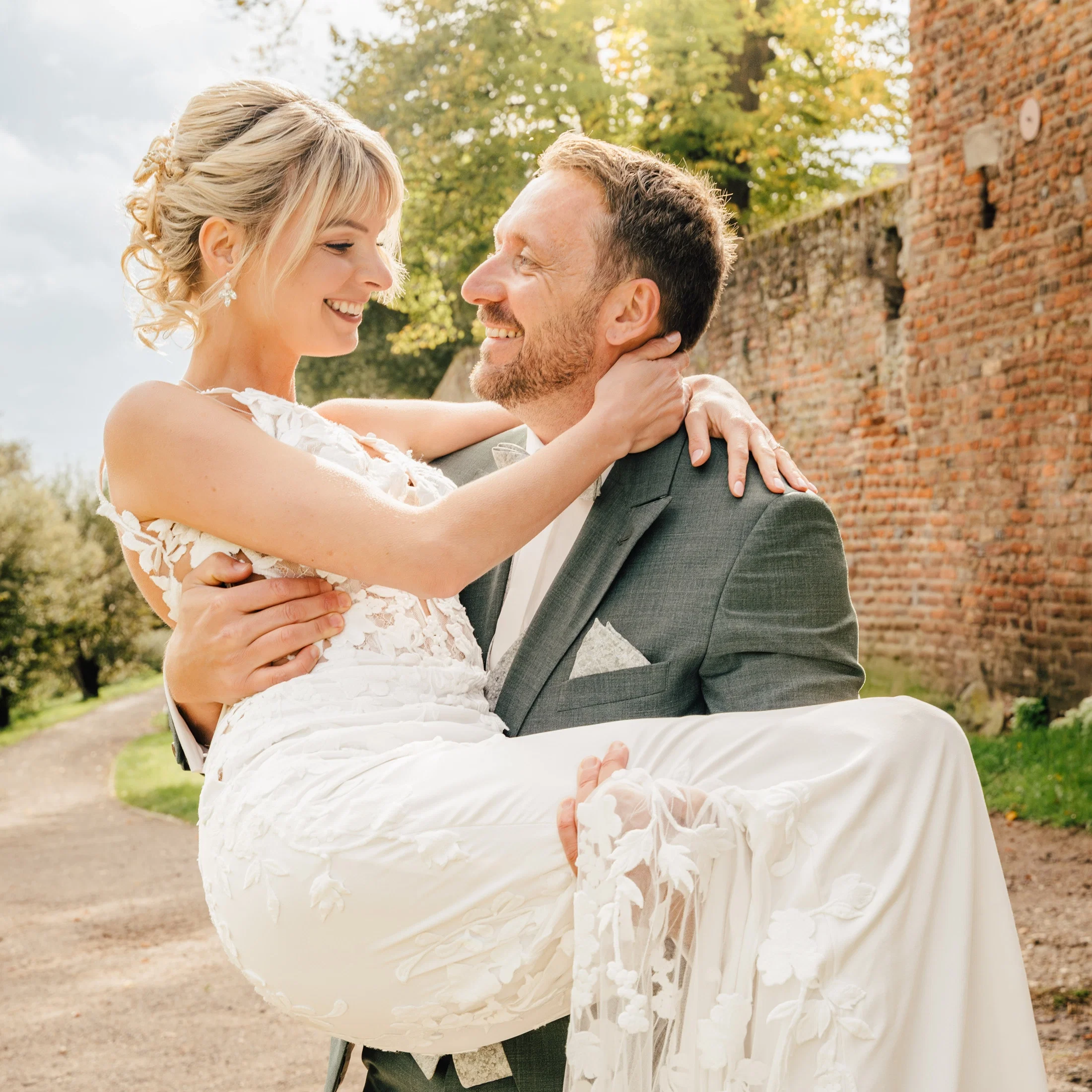 Brautpaar im Freien: Der Bräutigam trägt die Braut lächelnd auf den Armen, während sie sich verliebt ansehen, vor einer alten Backsteinmauer im warmen Sonnenlicht