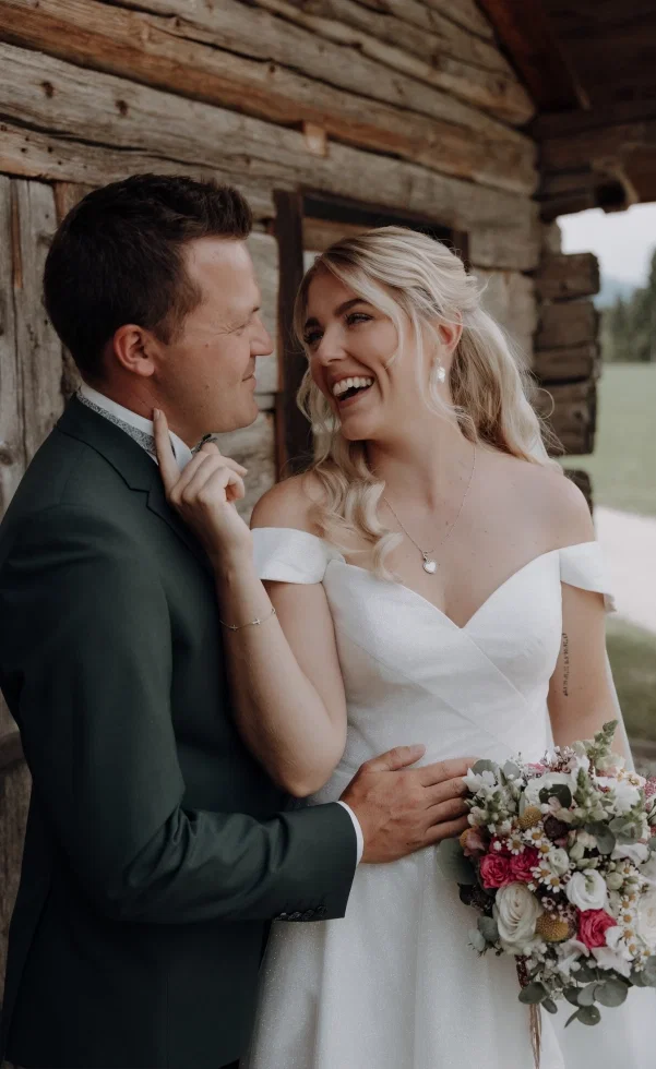 Romantisches Brautpaar an einer Holzhütte: Die Braut im schulterfreien Kleid lächelt ihren Bräutigam an und berührt sanft sein Gesicht, während sie einen Blumenstrauß hält