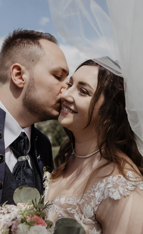 Zärtlicher Hochzeitsmoment: Der Bräutigam küsst die Braut liebevoll auf die Wange, während sie unter ihrem Schleier lächelnd in die Kamera blickt