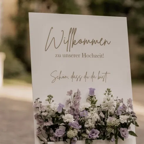 Willkommensschild zur Hochzeit mit zarter Blumenillustration in Weiß und Flieder, elegante Schrift mit der Aufschrift „Willkommen zu unserer Hochzeit – Schön, dass du da bist“