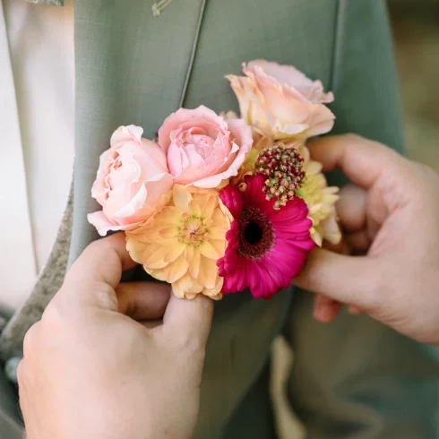 Hände stecken eine farbenfrohe Boutonniere mit Rosen, Dahlien und Gerbera an ein graues Anzugsrevers.
