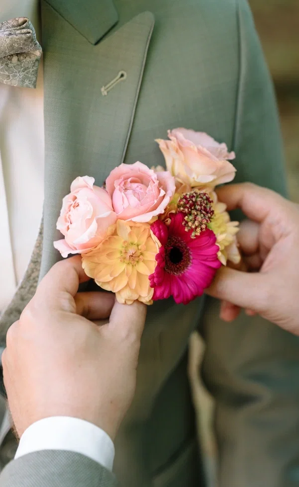 Detailaufnahme einer farbenfrohen Boutonniere mit Rosen, Dahlien und Gerbera, die an einem grünen Hochzeitsanzug befestigt wird.