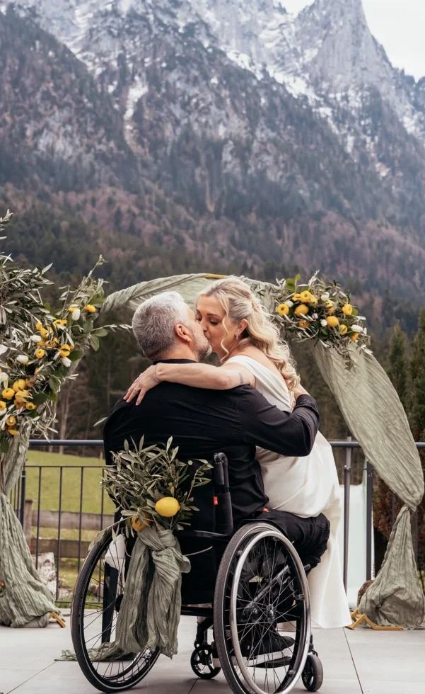 Romantischer Kuss bei barrierefreier Hochzeit: Braut und Bräutigam im Rollstuhl umarmen sich unter dekoriertem Traubogen vor Bergkulisse