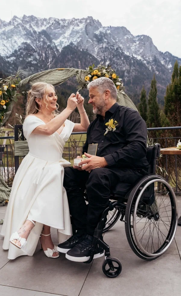 Emotionaler Moment bei barrierefreier Hochzeit: Die Braut steckt dem Bräutigam im Rollstuhl lachend den Ring an, während er sie glücklich ansieht