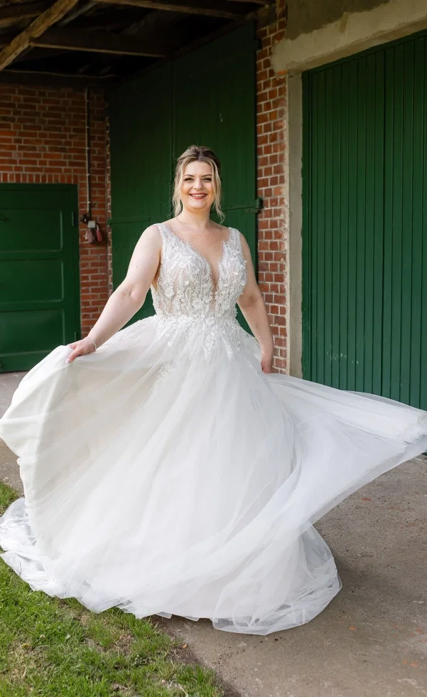 Strahlende Braut in einem luftigen Tüllkleid mit Spitzenoberteil, die vor grünen Scheunentoren ihr Kleid spielerisch schwingt.
