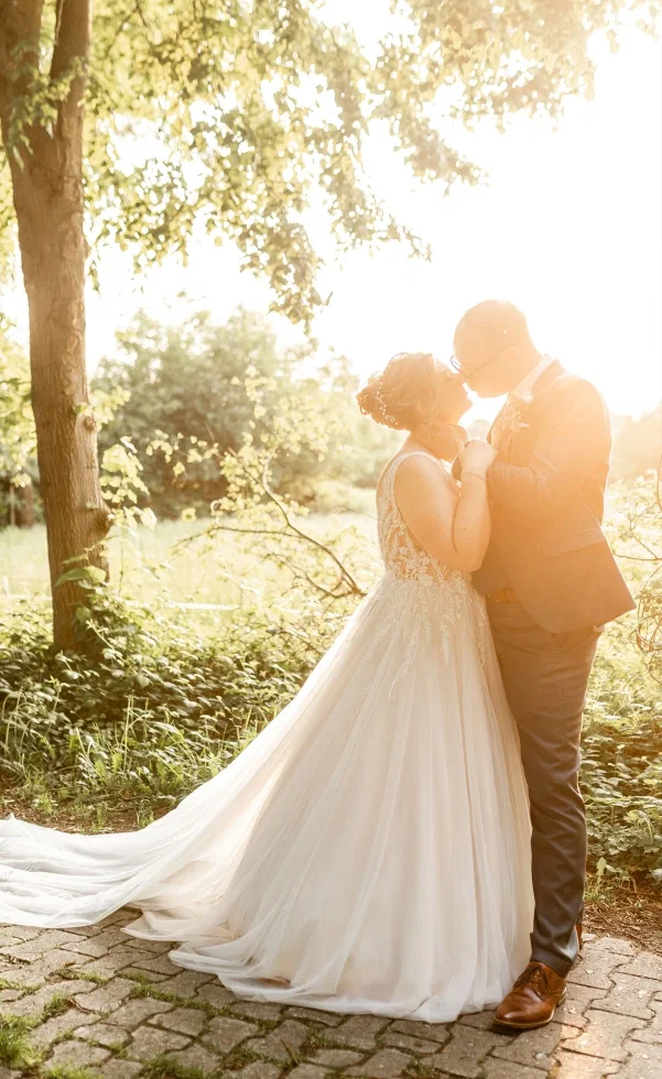 Brautpaar küsst sich im warmen Sonnenuntergang unter Bäumen, die Braut trägt ein fließendes Kleid mit Spitze und langer Schleppe.