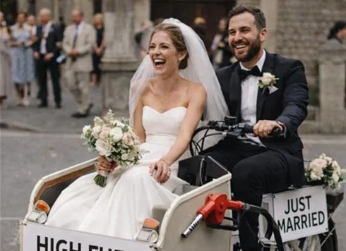 Frisch vermähltes Brautpaar fährt lachend in einem dekorierten Lastenfahrrad mit „Just married“-Schild, versehen mit einem humorvollen Hinweis auf hohe Spritpreise.