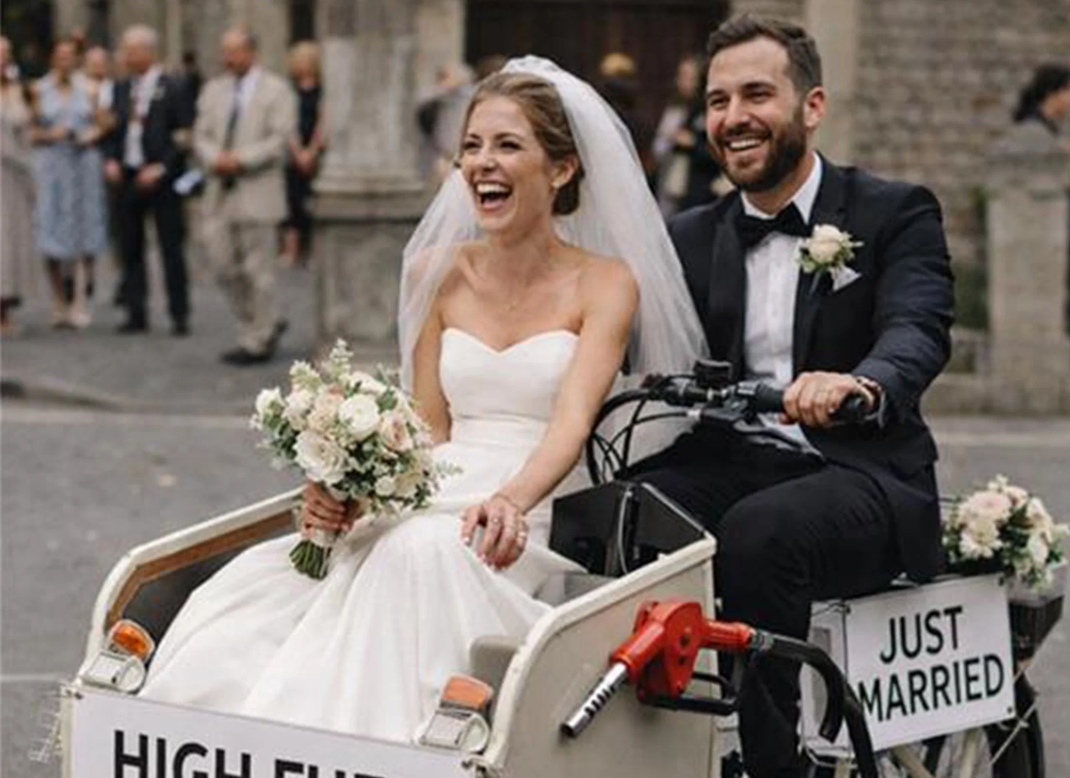 Frisch vermähltes Brautpaar fährt lachend in einem dekorierten Lastenfahrrad mit „Just married“-Schild, versehen mit einem humorvollen Hinweis auf hohe Spritpreise.