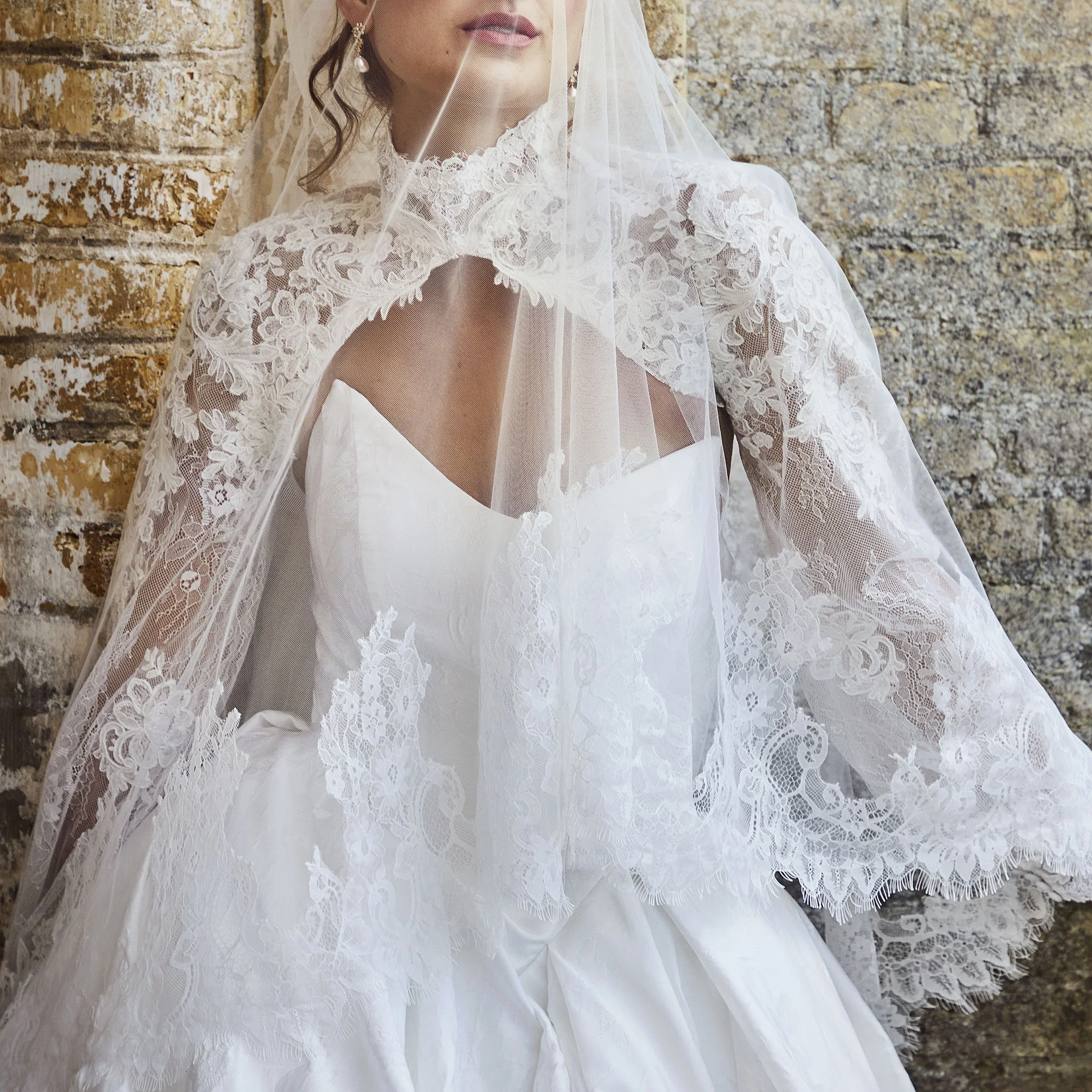 Braut in romantischem Brautkleid mit Spitzen-Cape und zartem Schleier vor rustikaler Steinwand