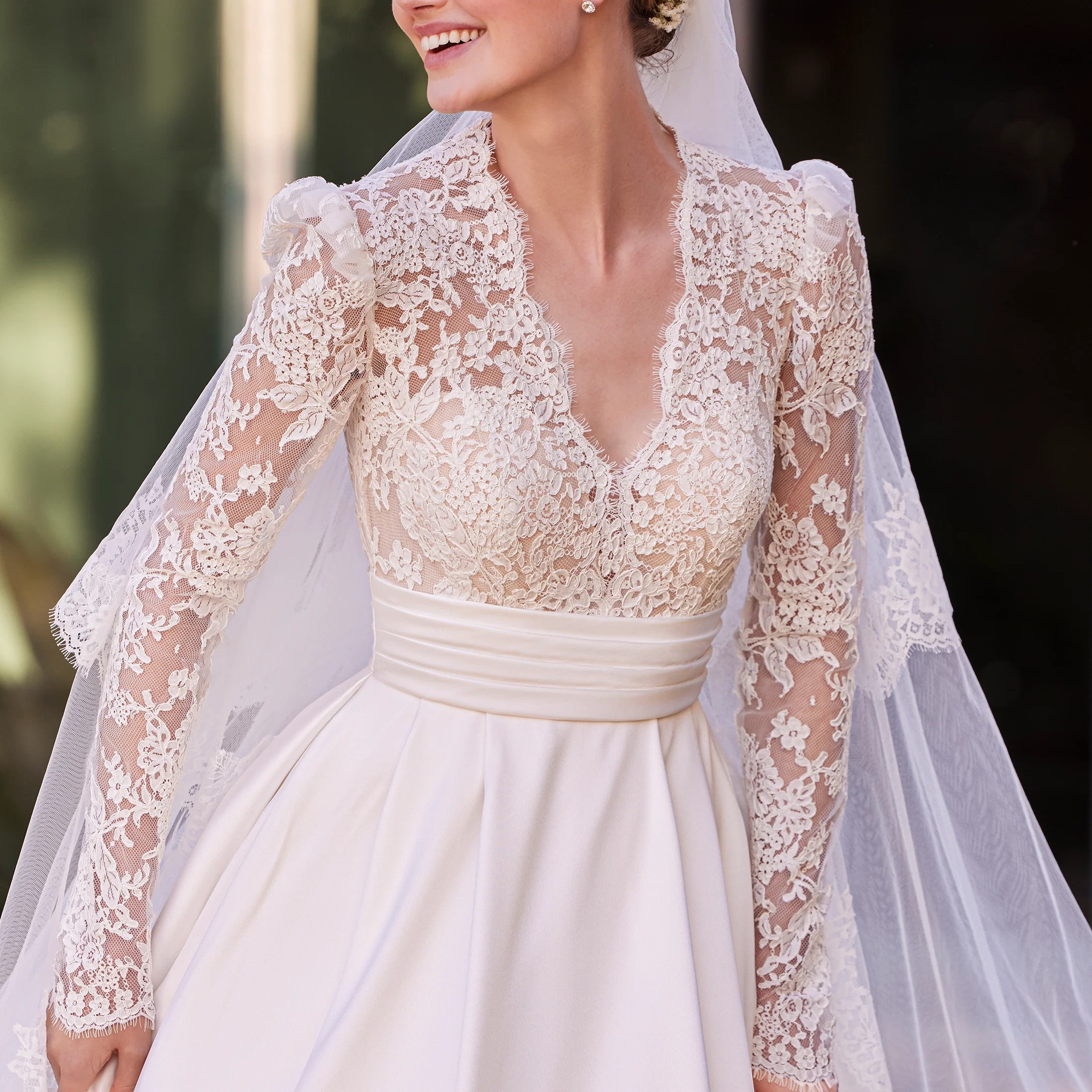 Braut in einem eleganten Brautkleid mit transparentem Spitzenoberteil, langen Ärmeln und floralen Stickereien, kombiniert mit einem fließenden Rock und zartem Schleier.