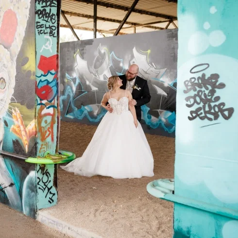 Brautpaar posiert innig in einer urbanen Industrial-Location mit Graffiti-Wänden, während das Brautkleid elegant im Kontrast zur rauen Umgebung steht