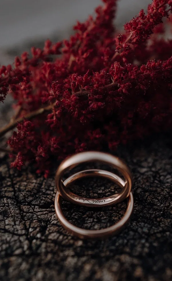 Zwei Eheringe liegen auf einer dunklen Oberfläche vor roten Blüten und wirken besonders stimmungsvoll inszeniert