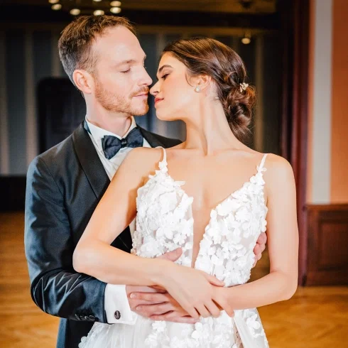 Ein elegantes Brautpaar tanzt eng umschlungen in einem festlichen Saal, während die Braut ein filigranes Spitzenkleid mit tiefem Ausschnitt trägt und der Bräutigam im schwarzen Anzug zärtlich zu ihr blickt.