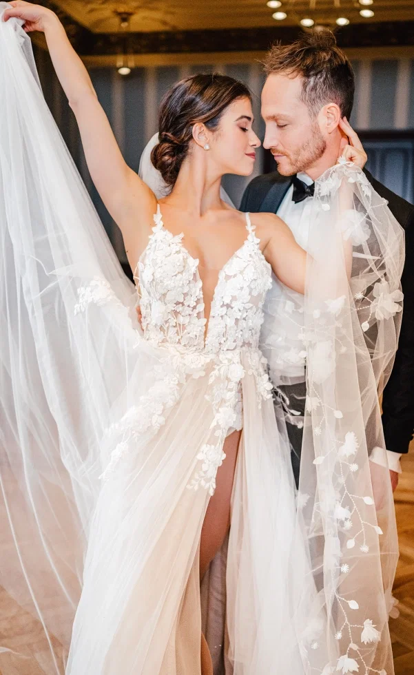 Die Braut hebt elegant ihren Schleier, während sie in einem detailreichen Spitzenkleid eng mit dem Bräutigam tanzt und beide sich liebevoll ansehen.