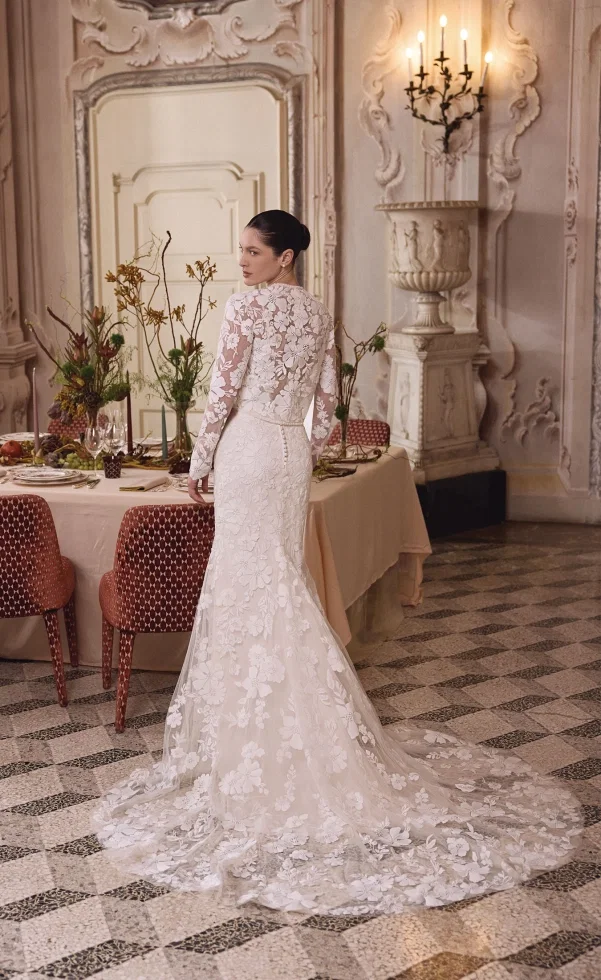Braut in einem figurbetonten Spitzen-Brautkleid mit langen Ärmeln und floralen Applikationen, kombiniert mit einer eleganten Schleppe, stehend in einem stilvollen, klassischen Interieur mit gedeckter Tafel.