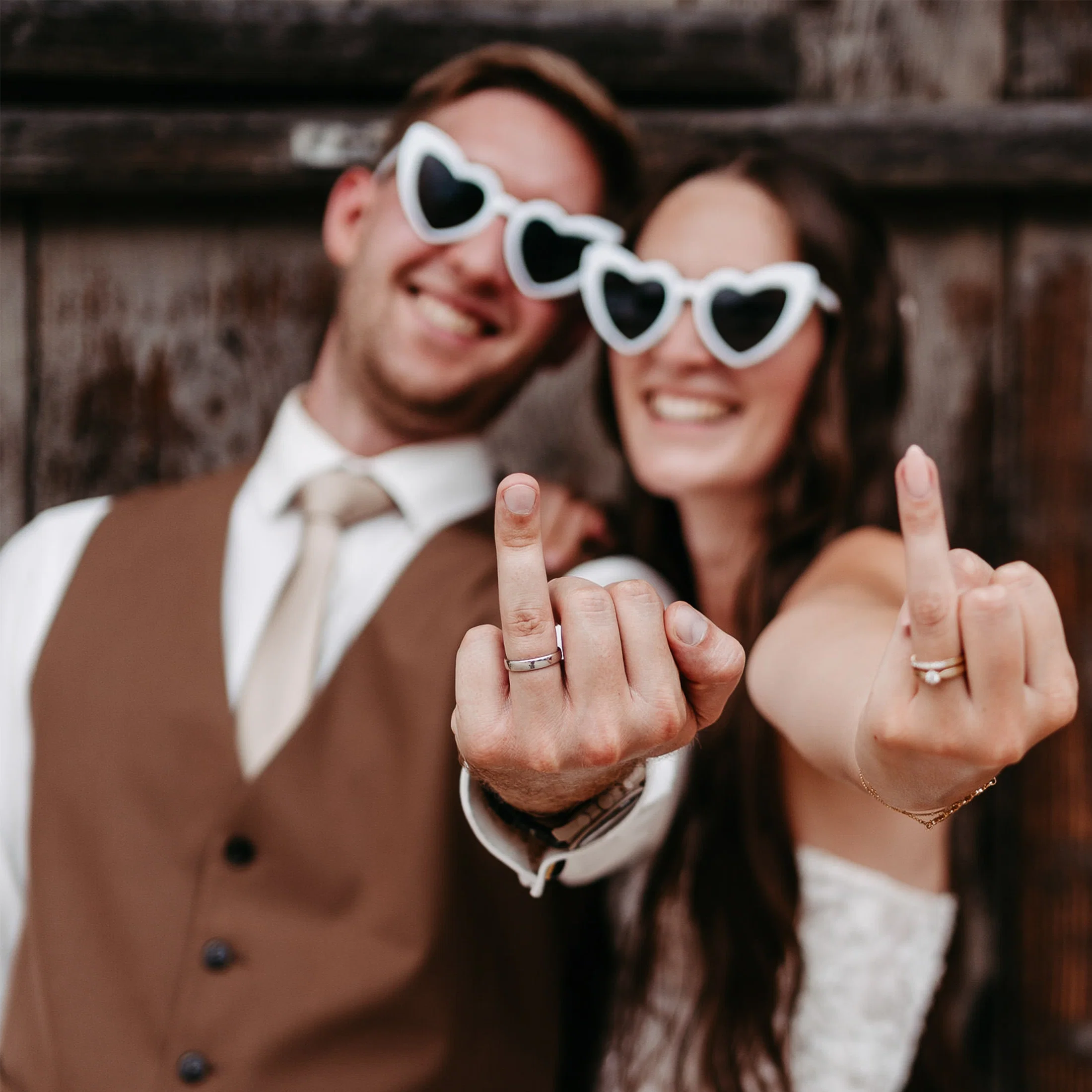 Frisch verheiratetes Paar mit Herz-Sonnenbrillen zeigt humorvoll den Mittelfinger, während ihre Eheringe im Vordergrund im Fokus stehen.