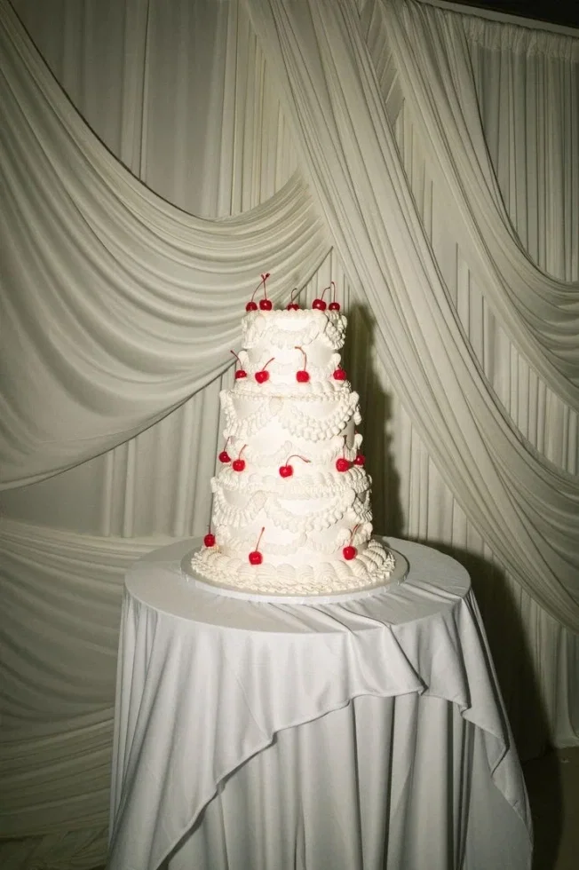 Mehrstöckige Hochzeitstorte in Cremeweiß mit kunstvollen Verzierungen, dekoriert mit roten Kirschen, präsentiert vor drapierten Stoffbahnen.