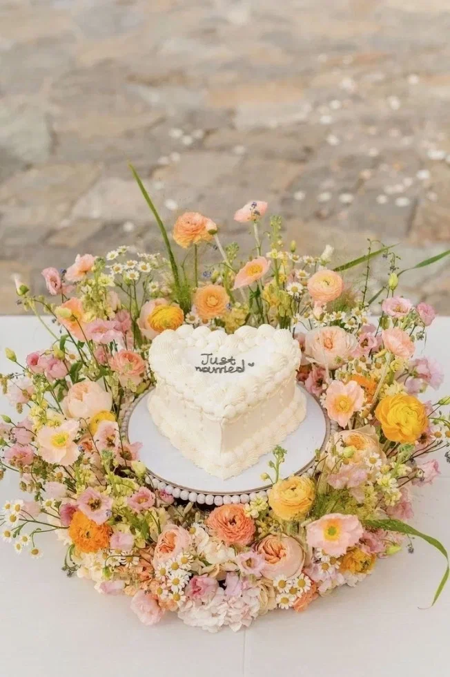 Herzförmige Hochzeitstorte in Cremeweiß mit „Just married“-Schriftzug, umgeben von einem üppigen Kranz aus zarten Blumen in Pastellfarben.