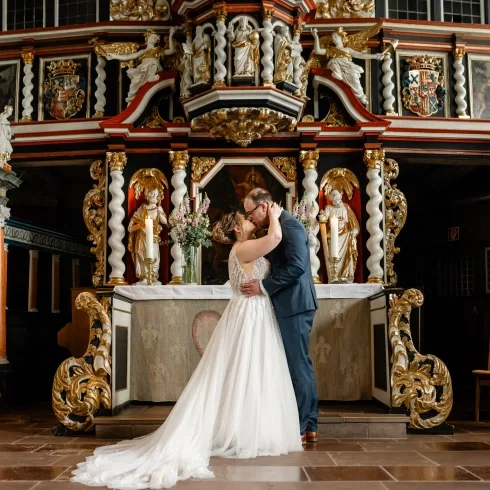 Brautpaar küsst sich vor einem prachtvollen barocken Altar in einer Kirche, die Braut trägt ein fließendes Tüllkleid mit Spitze, der Bräutigam einen dunklen Anzug.