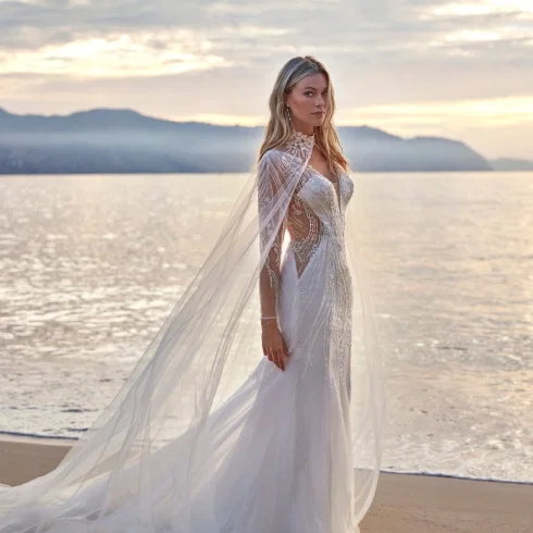 Brautkleid aus der Kollektion Deluxe by Amelie: Figurbetontes Meerjungfrauen-Kleid mit aufwendig perlensticktem, transparentem Oberteil in geschwungenen Musterlinien, kombiniert mit einem langen, fließenden Tüll-Cape mit besticktem Stehkragen. Das Cape reicht bodenlang und verleiht dem Gesamtlook eine dramatische, königliche Silhouette. Fotografiert bei Sonnenuntergang am Meer.