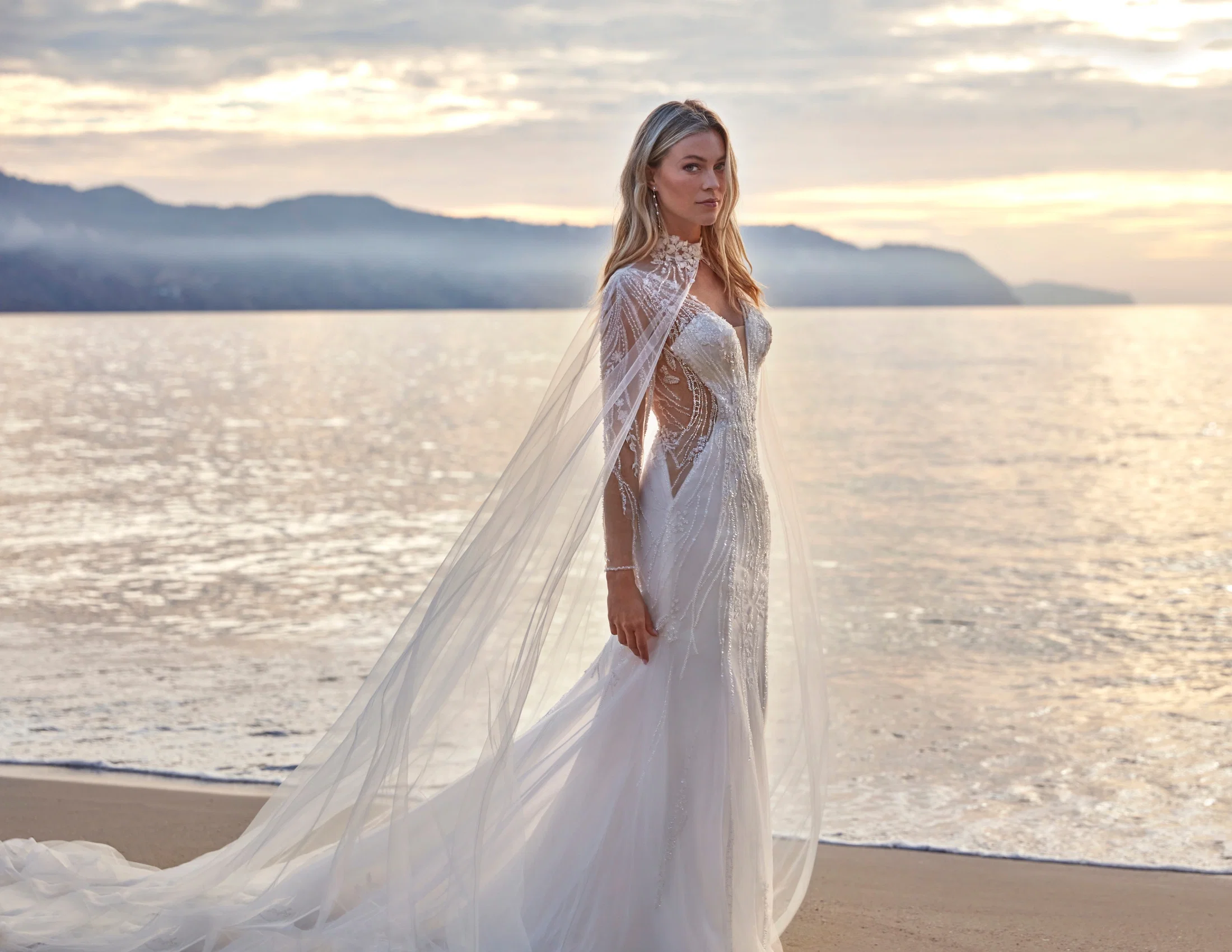 Brautkleid aus der Kollektion Deluxe by Amelie: Figurbetontes Meerjungfrauen-Kleid mit aufwendig perlensticktem, transparentem Oberteil in geschwungenen Musterlinien, kombiniert mit einem langen, fließenden Tüll-Cape mit besticktem Stehkragen. Das Cape reicht bodenlang und verleiht dem Gesamtlook eine dramatische, königliche Silhouette. Fotografiert bei Sonnenuntergang am Meer.