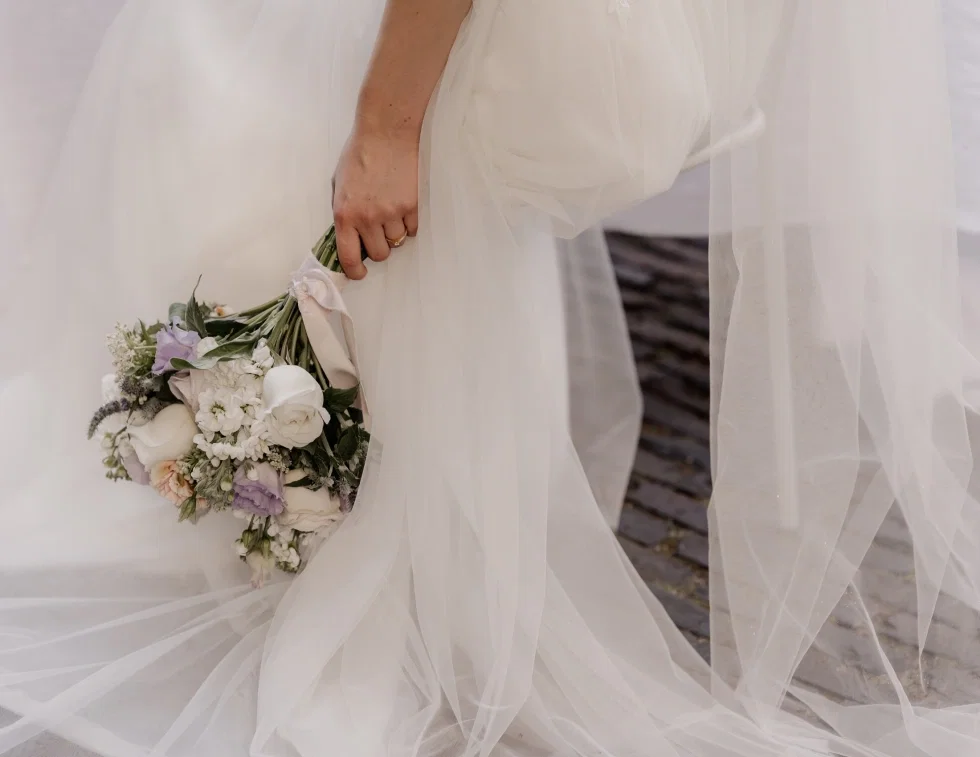 Detailaufnahme einer Braut im weißen Tüll-Hochzeitskleid, die mit einer Hand mit Goldring einen romantischen Brautstrauß aus weißen Rosen, lila Lisianthus, Pfingstrosen und zarten Wiesenblumen hält