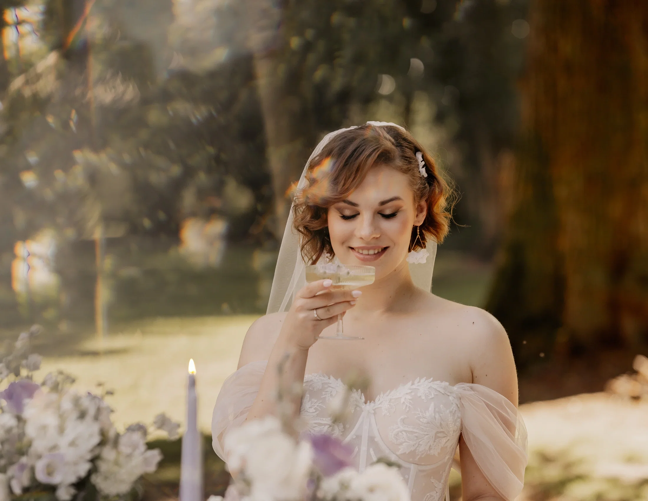 Strahlende Braut im bestickten Off-Shoulder-Hochzeitskleid mit Schleier hält lächelnd ein kleines Glas in der Hand, umgeben von warmem Sonnenlicht zwischen Bäumen und lila Kerzen im Vordergrund