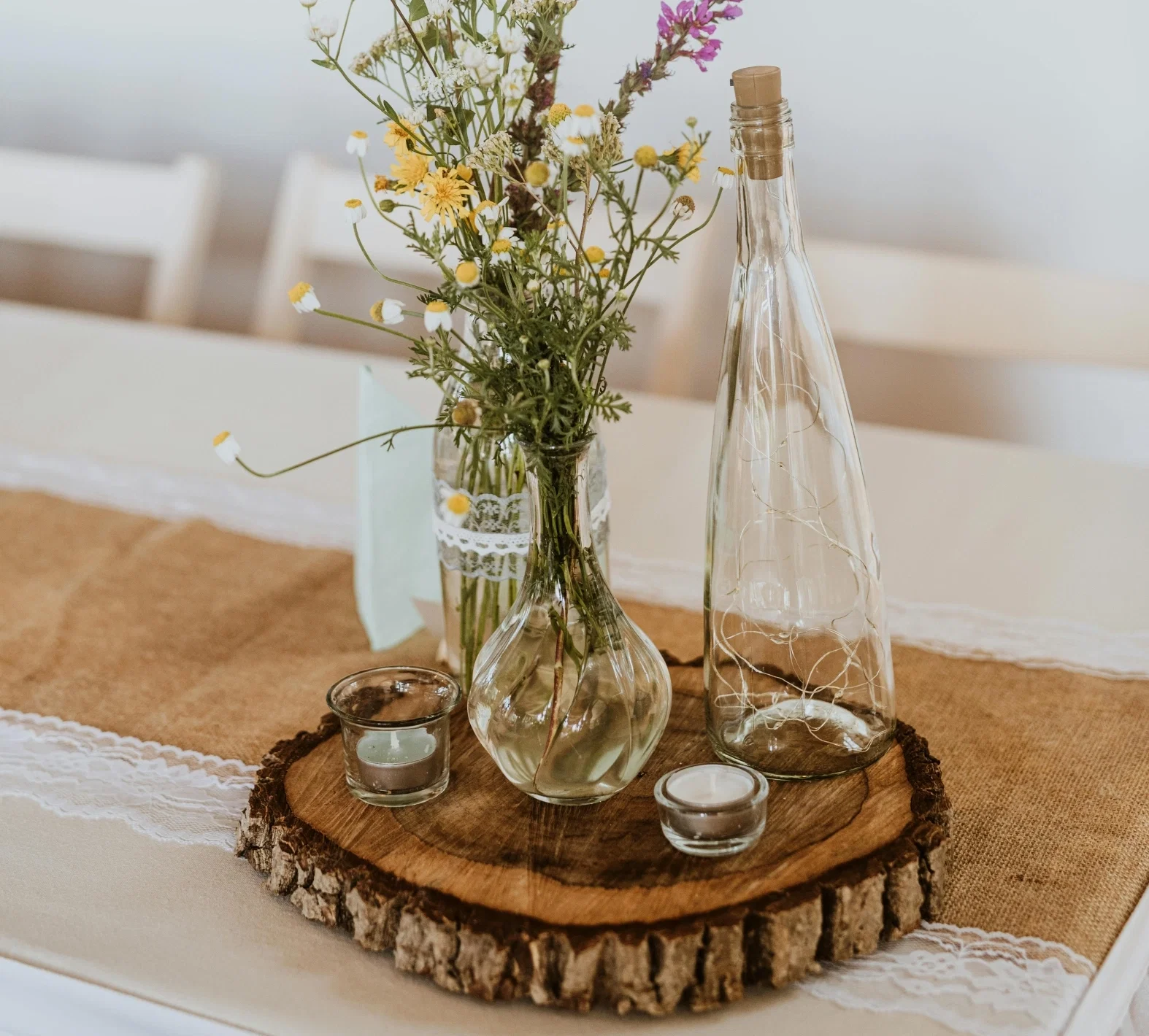 Rustikale Tischdeko mit Wildblumenstrauß in Glasvasen, Windlichtern und Deko-Flasche auf einer Baumscheibe