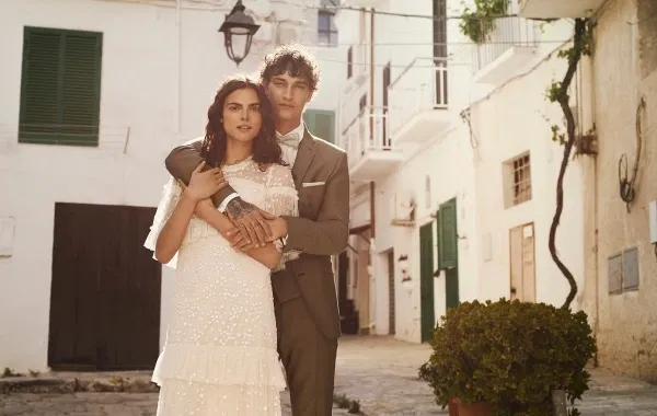 Elegantes Brautpaar in mediterraner Altstadt: Braut trägt ein modernes, fließendes Brautkleid mit feinen Stickereien, Bräutigam einen stilvollen beigen Hochzeitsanzug – romantisches Hochzeitsfoto bei natürlichem Tageslicht.