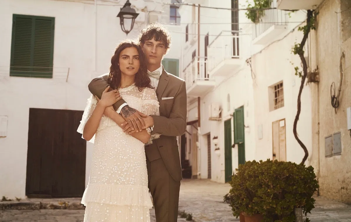 Elegantes Brautpaar in mediterraner Altstadt: Braut trägt ein modernes, fließendes Brautkleid mit feinen Stickereien, Bräutigam einen stilvollen beigen Hochzeitsanzug – romantisches Hochzeitsfoto bei natürlichem Tageslicht.